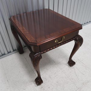 May include: A dark brown wooden side table with a rectangular top and a single drawer. The table features ornate, carved legs and a brass drawer pull. The table measures approximately 24 inches wide, 24 inches deep, and 24 inches high.