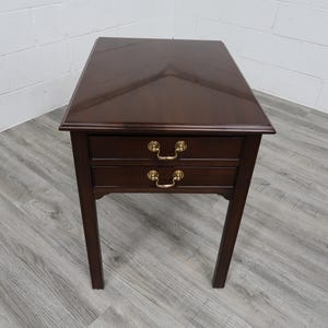 May include: A dark brown wooden end table featuring a smooth, polished rectangular top with a subtle V-shaped grain pattern. The table includes two stacked drawers, each equipped with two curved brass-toned metal handles, supported by four legs.
