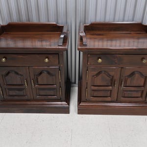 May include: Two matching dark brown wooden nightstands with a single drawer and two doors. Each nightstand has a decorative top edge and brass hardware.