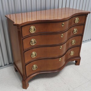 May include: A curved, wooden chest of drawers with five drawers and brass hardware. The chest is a rich brown color and has a slightly distressed finish.