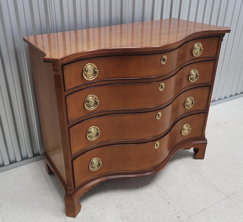 May include: A curved, wooden chest of drawers with five drawers and brass hardware. The chest is a rich brown color and has a slightly distressed finish.