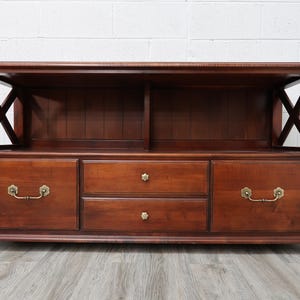 May include: A brown wooden media console featuring an open top shelf with X-shaped side supports, two side cabinets with brass-toned metal handles, and two central stacked drawers with small floral-shaped brass knobs, all resting on bun feet.