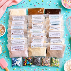 May include: A collection of baking mix pouches and small bags of colorful sprinkles arranged on a bright blue surface. The pouches contain various dry ingredients and include white labels with printed baking instructions and product names.