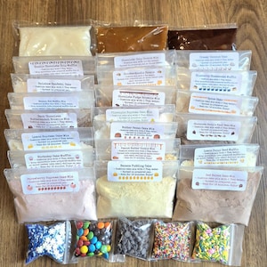 May include: An assortment of baking mixes in clear plastic bags, including cake, cookie, and muffin mixes. The bags are labeled with the names of the mixes, such as "Double Chocolate Chip Muffin" and "Strawberry Supreme Cake Mix". Sprinkles and chocolate chips are also visible.