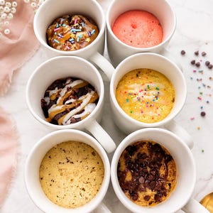 May include: Six white ceramic mugs arranged in two columns, each containing a different baked mug cake. The cakes feature various toppings including colorful sprinkles, chocolate chips, caramel drizzle, white icing, and a plain pink sponge cake.