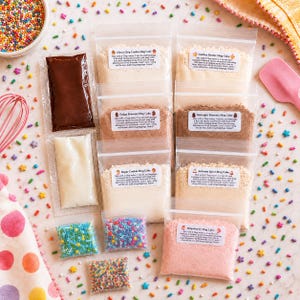 May include: A collection of seven clear plastic pouches containing various dry mug cake mixes, including Choco Chip, Golden Butter, Fudge Brownie, Midnight Brownie, Sugar Cookie, Autumn Spice, and Strawberry, plus three small bags of colorful sprinkles.
