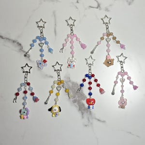 May include: Seven beaded keychains with star-shaped clasps. Each keychain features a different colour scheme of beads, charms, and decorative elements, including musical notes and cartoon characters. The keychains are displayed on a white marble surface.