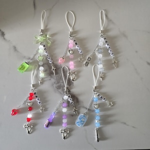May include: A collection of six beaded phone charms with various designs. Each charm features a white loop, silver chain, and decorative beads in colors like pink, red, and blue. Some charms include text like "TWICE" and "NCT127".
