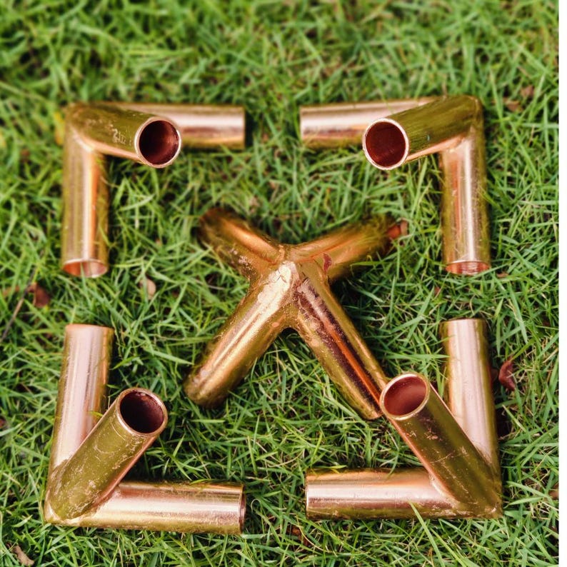 May include: Four copper pipe elbow fittings arranged in a square formation surrounding a central copper pipe cross fitting. Each metallic component features a hollow cylindrical design with a polished finish, showcasing the distinct reddish-brown hue.