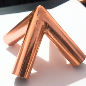 May include: A three-way copper pipe fitting with a metallic, reddish-brown finish. The connector features three cylindrical arms joined at a central point, forming a tripod-like structure with smooth, polished surfaces and visible seams at the joints.