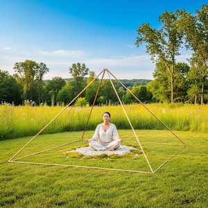 May include: A large, hollow, copper-colored metal pyramid frame constructed from thin, straight rods. The structure is positioned on a patch of green grass, surrounding a circular area of white stones and moss where a person sits in meditation.