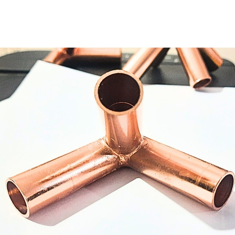 May include: A three-way copper pipe fitting with a metallic, reddish-brown finish. The connector features three cylindrical openings arranged at right angles to each other, designed to join pipes at a corner junction in a plumbing system.