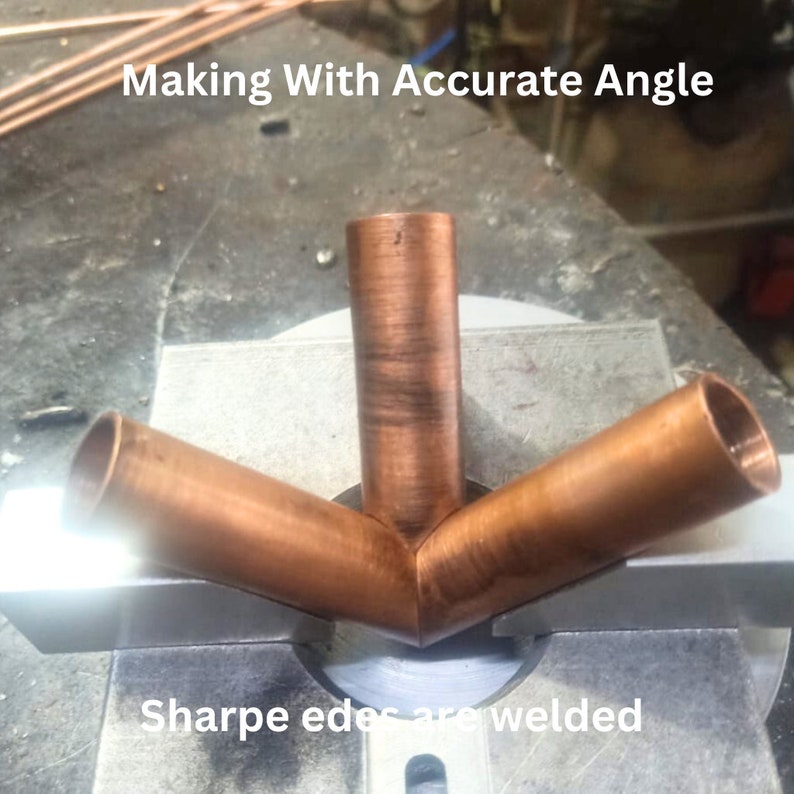 May include: A copper pipe fitting with three cylindrical branches joined at a central point, featuring the text Making With Accurate Angle and Sharpe edes are welded. The metallic surface shows a reddish-brown tone with visible texture.