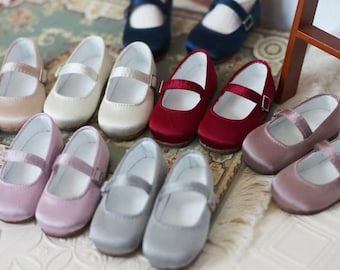 May include: A collection of satin Mary Jane style shoes in various colors, including cream, pink, red, and gray. Each shoe features a strap with a buckle closure. The shoes are arranged on a patterned surface.