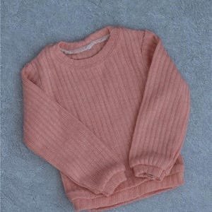 May include: A coral-colored sweater with a ribbed texture. The long-sleeved top has a crew neck and is made of a soft, knitted material. The sweater is laid flat on a gray surface.