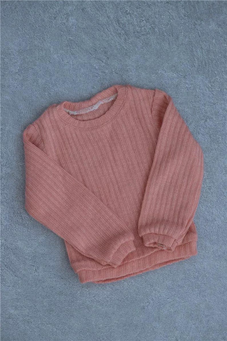 May include: A coral-colored sweater with a ribbed texture. The long-sleeved top has a crew neck and is made of a soft, knitted material. The sweater is laid flat on a gray surface.