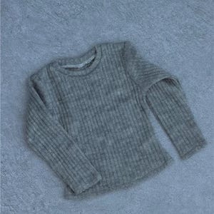 May include: A gray, ribbed knit long-sleeve shirt. The shirt has a crew neck and is made of a soft, textured material. The garment is displayed on a gray surface.