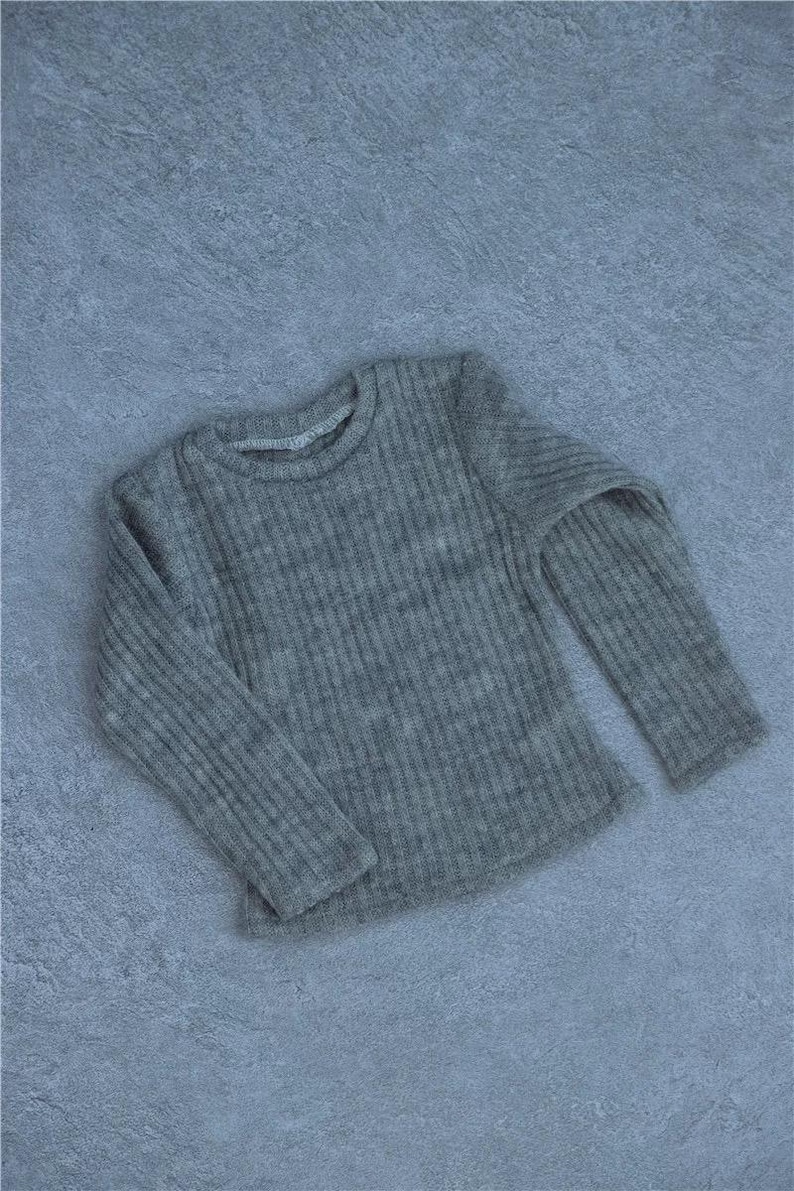 May include: A gray, ribbed knit long-sleeve shirt. The shirt has a crew neck and is made of a soft, textured material. The garment is displayed on a gray surface.