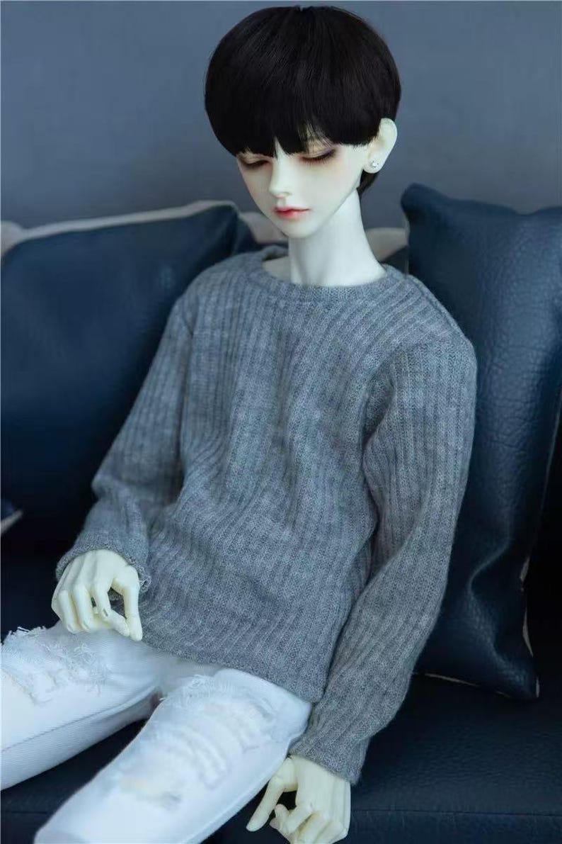 May include: A doll with a pale complexion, black hair, and a gray sweater. The doll is wearing white ripped jeans. The doll is sitting on a blue surface.