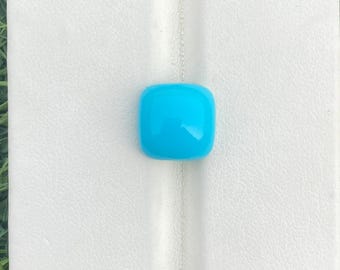 May include: A vibrant, square-shaped, turquoise gemstone. The polished stone is set on a white leather surface, with a green grassy background. The gemstone's smooth, rounded edges and bright color make it a striking focal point.