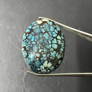 May include: Oval turquoise gemstone with a dark matrix. The stone has a mosaic pattern of blue and green hues, held by tweezers. The gemstone is likely used for jewellery making or collecting.