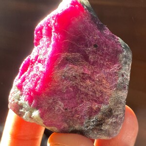 Peut inclure: Une pierre précieuse brute et non polie présentant un centre magenta-rose vibrant et saturé qui se transforme en inclusions minérales gris foncé et noires opaques sur les bords, avec une texture cristalline rugueuse et une forme irrégulière.