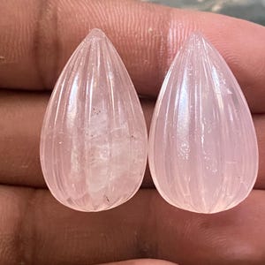 Puede incluir: Dos gemas de cuarzo rosa talladas en forma de lágrima. Cada gema tiene ranuras verticales y una superficie pulida, mostrando un suave tono rosado. Las piedras se sostienen en una mano, mostrando su calidad translúcida y belleza natural.