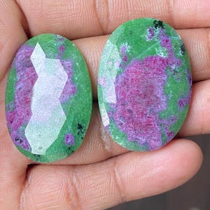 Può includere: Due pietre preziose ovali con superfici sfaccettate. Le pietre presentano una base verde vibrante con un motivo irregolare prominente di tonalità rosa e rosse intense. Sono visibili piccole inclusioni nere.