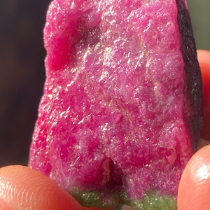 以下が含まれることがあります： 鮮やかで彩度の高いマゼンタピンクの結晶面を持つ、未研磨の原石。表面は粗く質感があり、石の底部には対照的なマットで落ち着いた緑色の鉱物素材の帯が見られます。