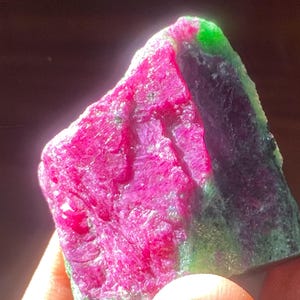 以下が含まれることがあります： 鮮やかなマゼンタピンクの中央部と、深緑色および濃い紫色の結晶質の縁が対照的な、未研磨の原石標本。石の表面は粗く質感があり、自然なひび割れや光を通す半透明の部分が見られます。