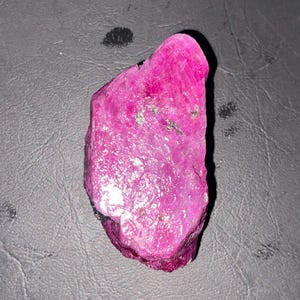 Puede incluir: Una piedra preciosa de rubí en bruto y sin pulir con un color magenta rosado vibrante y saturado. La piedra presenta una forma irregular y dentada con una superficie cristalina texturizada que refleja la luz, mostrando facetas naturales.