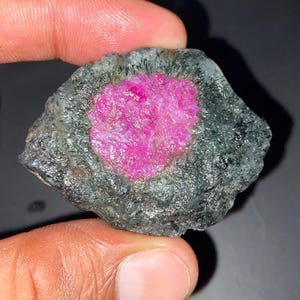 May include: A raw, uncut gemstone featuring a vibrant, textured magenta-pink centre surrounded by a dark, mottled grey-green outer layer. The stone has an irregular, organic shape with a rough, crystalline surface texture throughout the specimen.