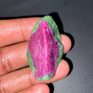 Puede incluir: Una piedra preciosa en bruto y sin pulir que presenta un centro de color magenta rosado intenso rodeado por un borde mineral de color verde claro texturizado. La piedra tiene una forma irregular y alargada con una superficie rugosa.