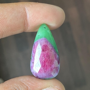 May include: A teardrop-shaped gemstone with a faceted, deep pink lower half and a smooth, green upper portion. The gem is held between two fingers, showcasing its polished surface and color contrast. The gem is likely a ruby.