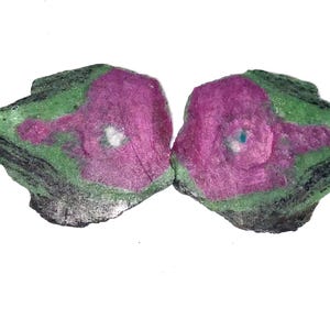 Könnte beinhalten: Zwei rohe Zoisit-Mineralspezimen mit leuchtend magentafarbenen Zentren, die von gesprenkelten grünen und schwarzen Außenschichten umgeben sind. Die Steine weisen unregelmäßige, gezackte Kanten und eine raue, unpolierte Textur mit sichtbaren Strukturen auf.