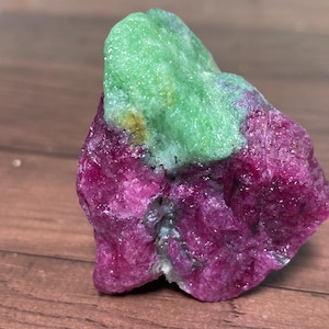 Puede incluir: Un espécimen mineral en bruto y sin pulir que presenta una sección de rubí de color magenta vibrante fusionada con un cristal de zoisita verde brillante y texturizado. La piedra muestra una forma natural e irregular con superficies cristalinas.