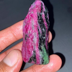 Puede incluir: Un espécimen mineral en bruto y alargado que presenta una combinación vibrante de secciones cristalinas de color rosa magenta intenso, negro opaco y verde brillante. La superficie tiene una textura natural rugosa con variaciones de color.