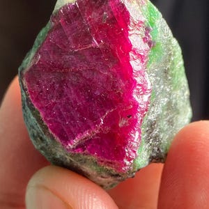 May include: A raw, unpolished mineral specimen featuring a vibrant, deep magenta-red crystalline centre embedded within a rough, textured matrix of opaque, mottled green and grey stone with natural jagged edges and a translucent, crystalline surface.