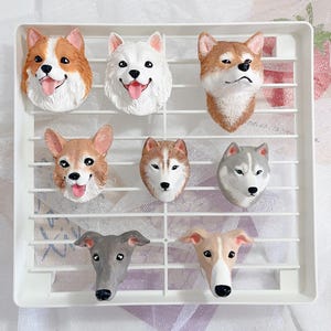 Puede incluir: Colección de imanes con forma de cabeza de perro, que representan varias razas como Corgis y Huskies. Los imanes están dispuestos en una rejilla blanca, mostrando sus detalles y colores realistas.