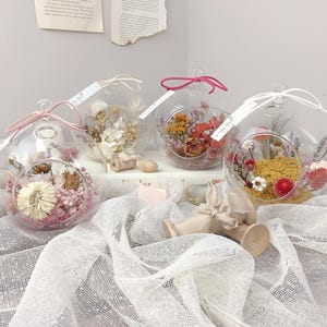 May include: Four spherical glass terrariums, each filled with dried flowers in shades of pink, yellow, and orange. The terrariums have a loop for hanging. White tags are attached. The terrariums are displayed on a white surface.