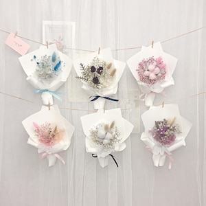 May include: Six miniature dried flower bouquets, each wrapped in white paper and tied with ribbons. The bouquets display a variety of colours, including blue, pink, and purple, and are hung from a string with small wooden pegs.