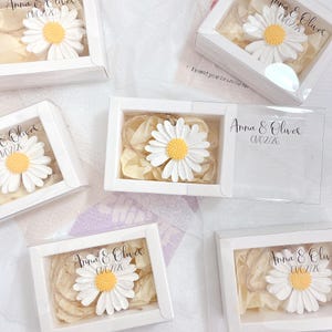 May include: White favour boxes with clear lids, each containing a small white daisy with a yellow centre. The boxes are decorated with the text "Anna & Oliver" and the date "01/02/26".