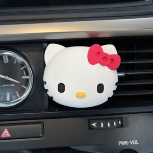 Puede incluir: Ambientador de coche Hello Kitty blanco con un lazo rojo. El ambientador está sujeto a una rejilla de ventilación del coche.