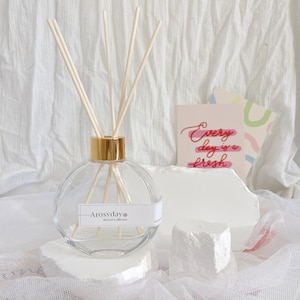 May include: A clear glass diffuser bottle with a gold lid and wooden diffuser sticks. The bottle is sitting on a white surface with a small white rock. The bottle has a label that says "Arosyday" with the text "Roses & Minerals" underneath.