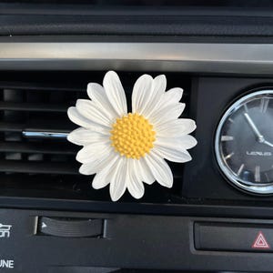 以下が含まれることがあります： 黄色の芯を持つ白いデイジーの車の芳香剤。芳香剤は車の通気口に取り付けられています。