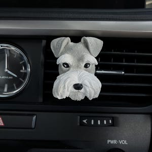 May include: A grey and white miniature schnauzer dog car air freshener. The air freshener is made of plastic and has a realistic design. It is attached to a car's air vent.
