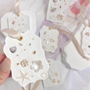 Pode incluir: Etiquetas de presente brancas decoradas com uma variedade de conchas e pérolas. As etiquetas são amarradas com uma fita branca.
