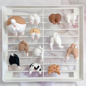 May include: A collection of 12 ceramic animal figurines, each depicting the rear end of a different animal, including a cat, dog, and rabbit. The figurines are painted in various colours, including brown, white, black, and orange. They are displayed on a white grid.