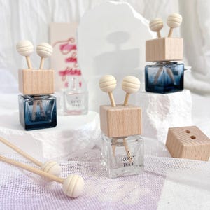 Könnte beinhalten: Vier gläserne Auto-Lufterfrischer mit Holzdeckeln und Holzstäbchen. Die Flaschen sind klar und enthalten eine blaue Flüssigkeit. Die Holzdeckel haben eine kleine runde Holzperle oben auf jedem Stäbchen. Die Flaschen sind mit "A Rosy Day" beschriftet.