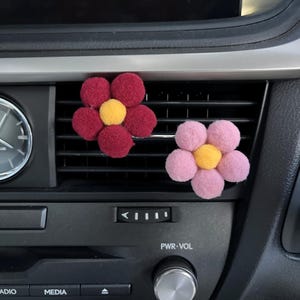 May include: Two fluffy flower-shaped car air fresheners. One is red with a yellow centre and the other is pink with a yellow centre. They are attached to a car's air vent.
